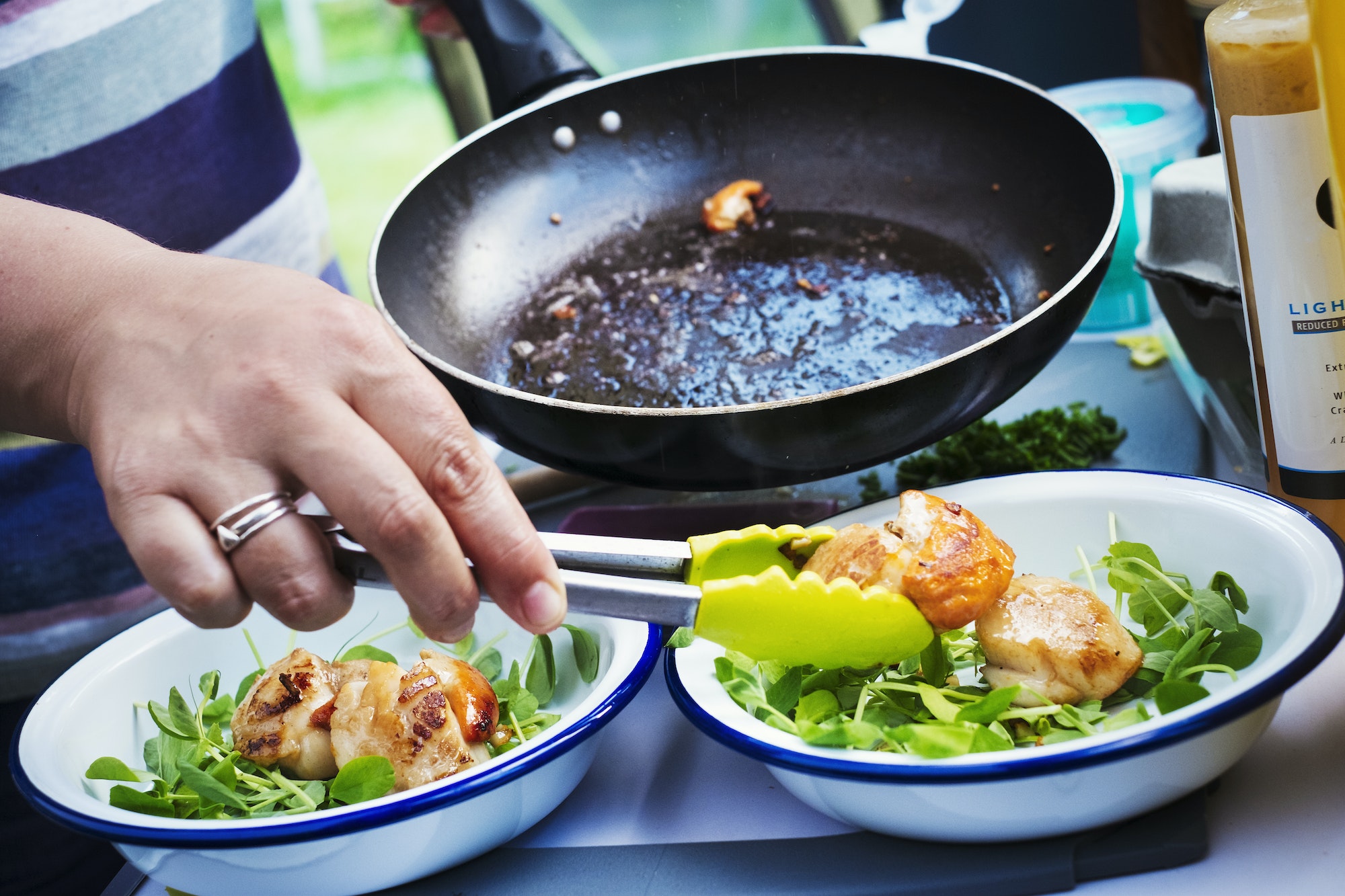 Woman standing at a camping stove, preparing two plates with fried scallops and fresh herbs.